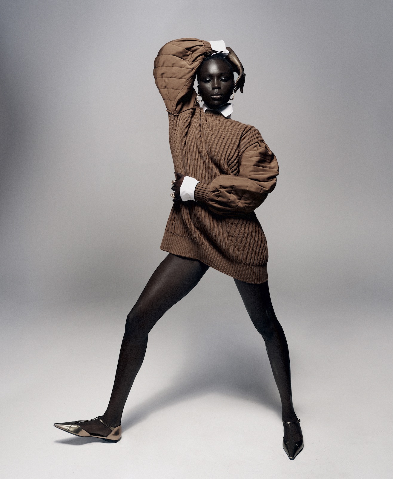A striking, dark-skinned model in a brown knit sweater and black tights poses against a grey backdrop in a fashion-forward editorial shot.