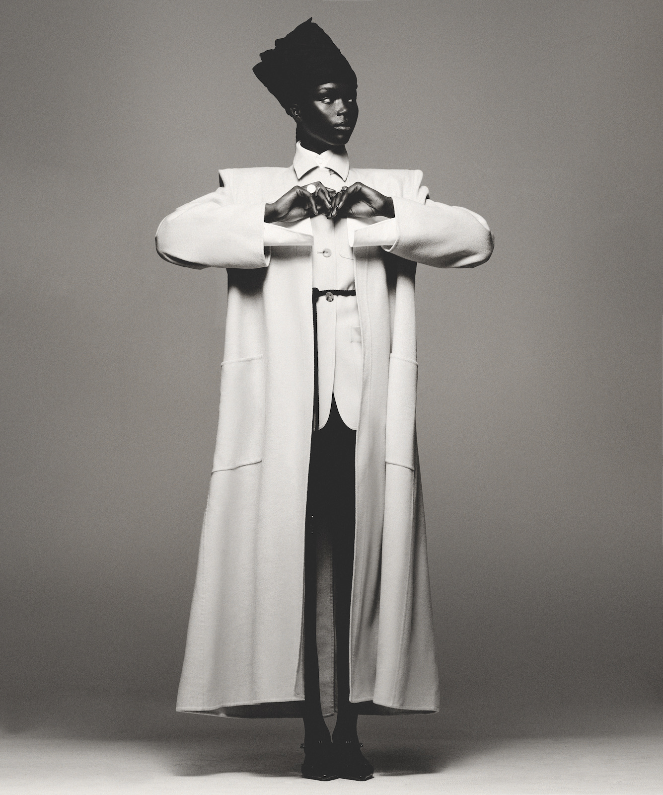 Fashionable person poses in studio wearing long white coat with dark top, pants, shoes, and tall cap against a gray background.
