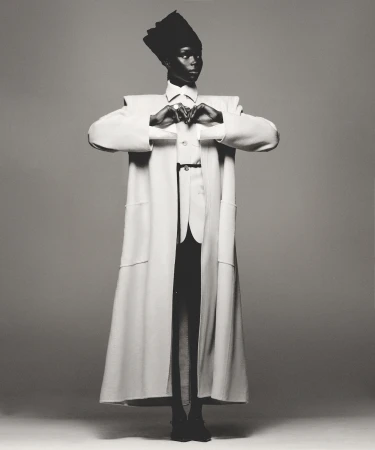 Fashionable person poses in studio wearing long white coat with dark top, pants, shoes, and tall cap against a gray background.