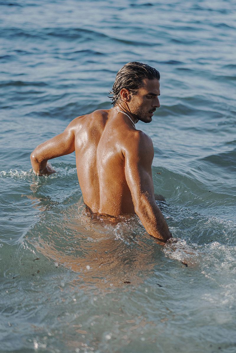 Man with dark hair and tan skin stands in blue ocean water, his back to the camera, looking off to the side with wet hair.