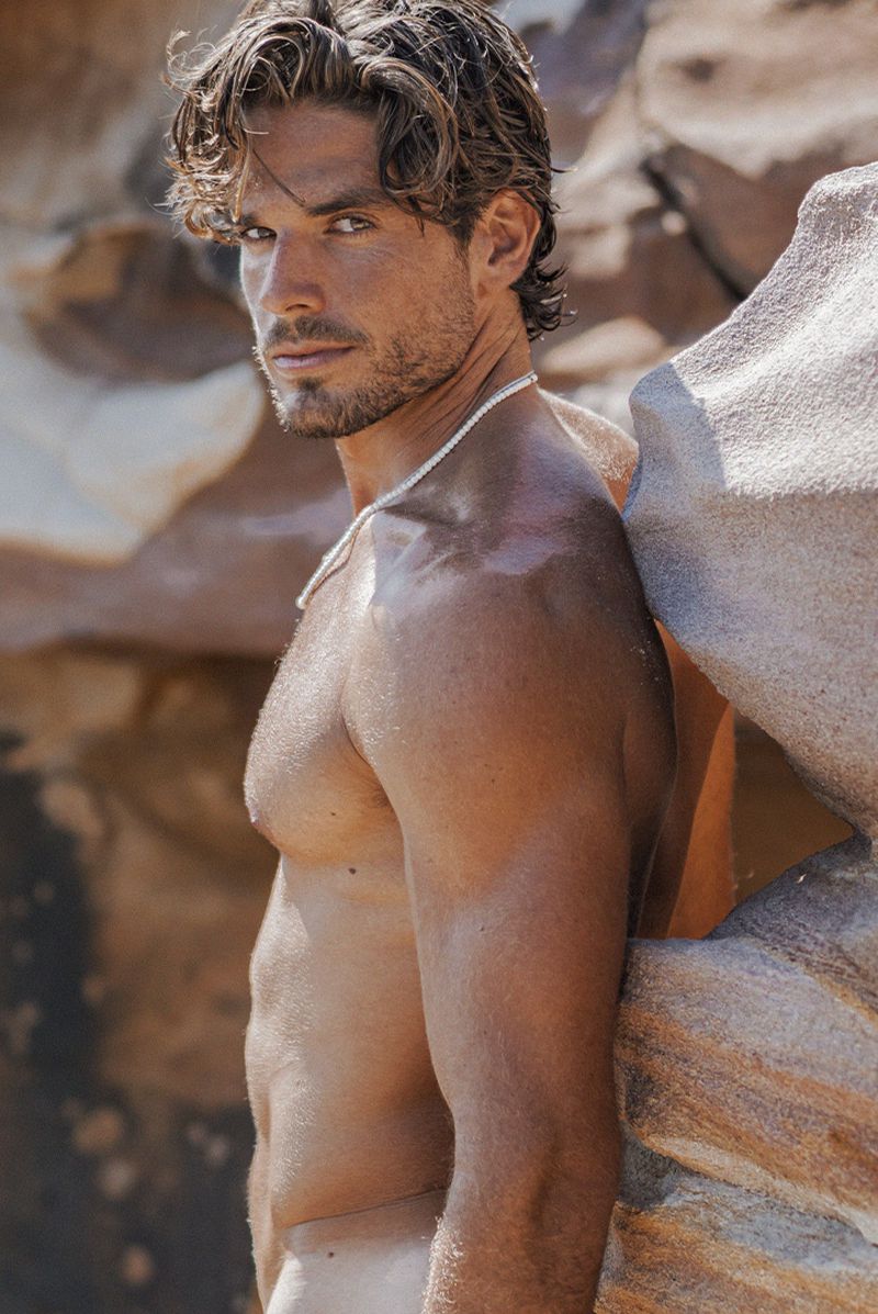 Portrait of a shirtless adult male with dark hair leaning against a weathered rock formation outdoors, looking at the camera.