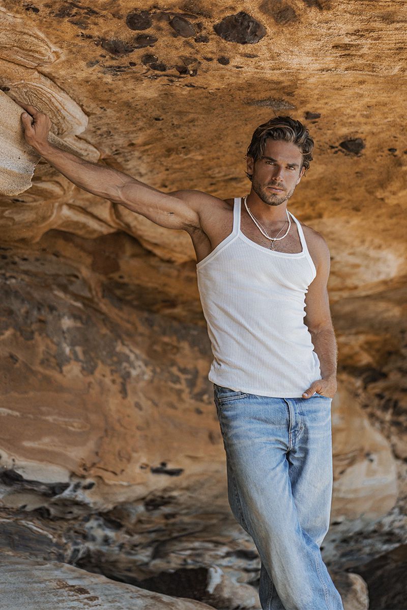 Adult male in a white tank top and jeans poses against a rock formation with golden brown hues in an outdoor setting.
