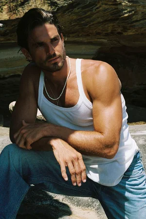 Man wearing a white ribbed tank top and blue jeans crouching near rocky terrain outdoors
