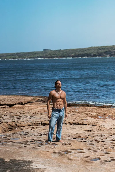 Shirtless man wearing blue jeans standing on a rocky coastal shore looking upward toward an ocean horizon.