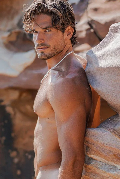 Shirtless man with dark curly hair leaning against a rocky sandstone wall outdoors in bright daylight