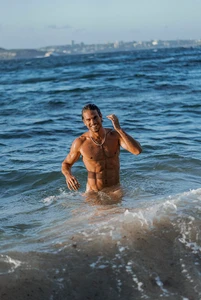 Shirtless man with wet hair smiling while standing in ocean waves near a shoreline