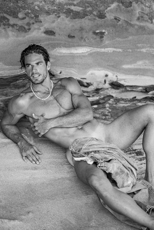 Monochrome image of a shirtless man lying against a rocky textured background with denim jeans draped near his waist.