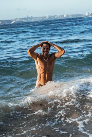 Shirtless man standing in ocean water holding his hands behind his head with waves breaking in the foreground