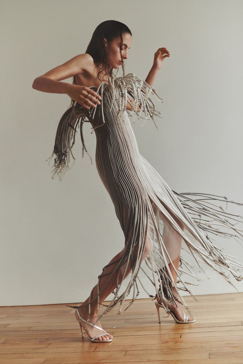 A slender woman in a flowing silver dress with fringed details strikes a dynamic pose on a wooden floor against a neutral background.