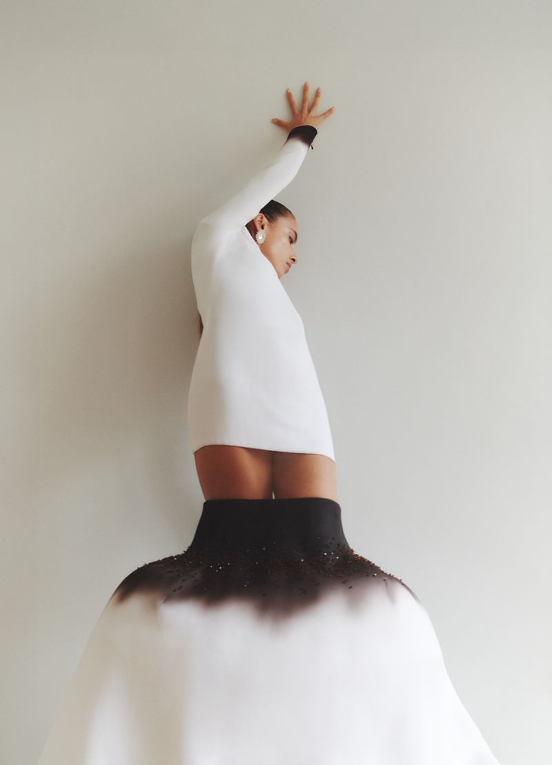 Woman in white dress with black details poses in front of a plain wall, hand up, shot in portrait orientation and eye-level angle.