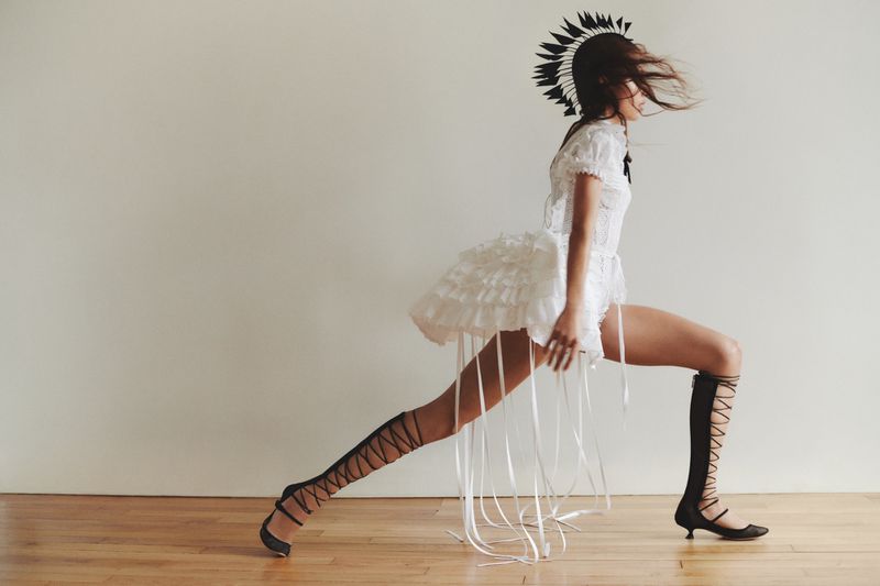 Side view of a woman in a white dress with a feathered headdress and lace-up knee-high boots striking a pose in a photo studio.