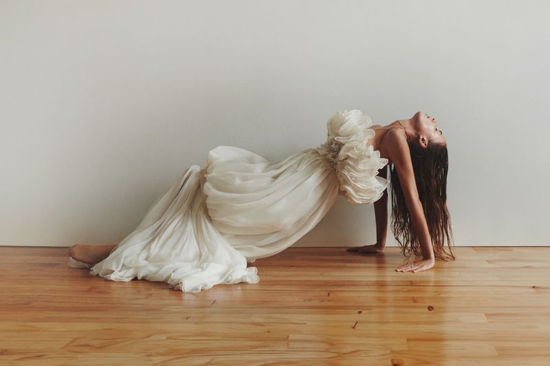 Woman in a flowing white dress arches backward on a wood floor, showcasing an open back and ethereal fabric against a neutral backdrop.
