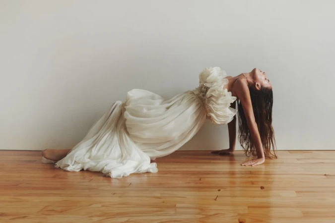 Woman in a flowing white dress arches backward on a wood floor, showcasing an open back and ethereal fabric against a neutral backdrop.