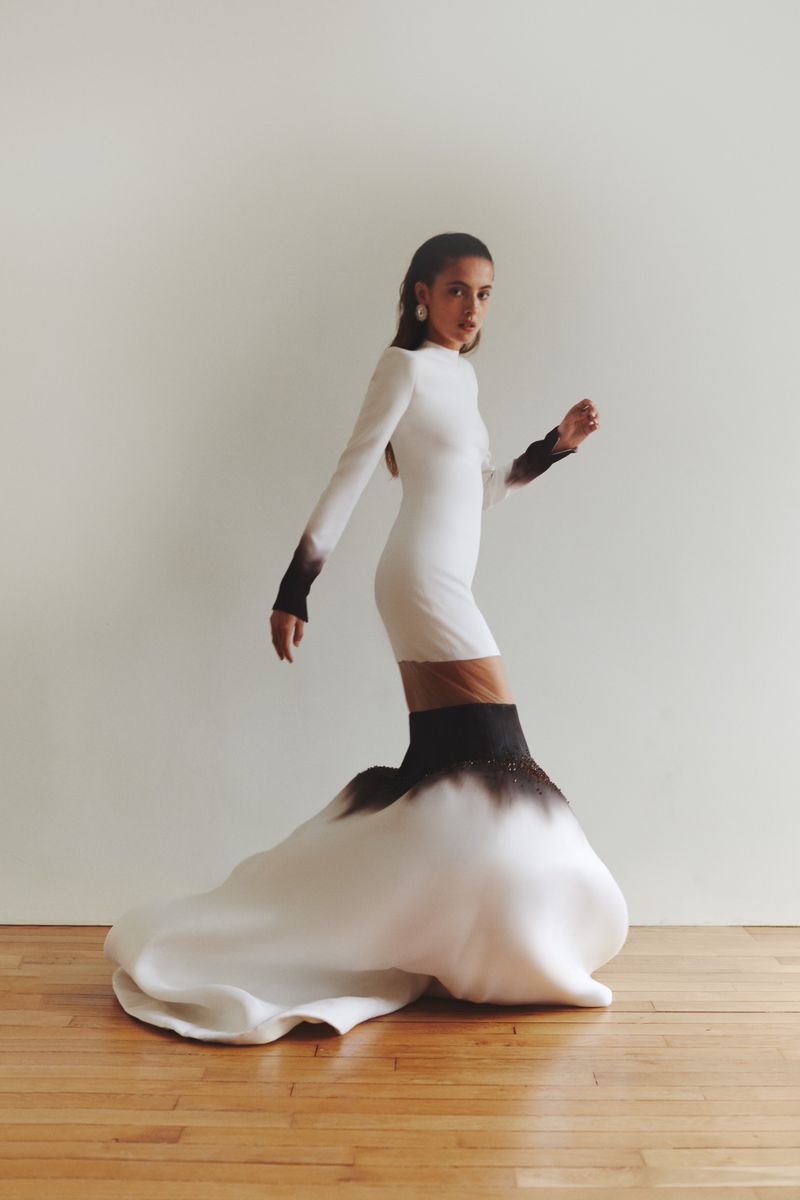 A woman posing in a studio wearing a white dress with long sleeves and black ombre detailing at the cuffs and bottom hem.
