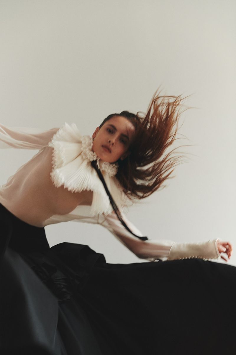 A woman with flowing auburn hair, posing dynamically in a sheer white blouse with a large ruffled collar and a long black skirt.