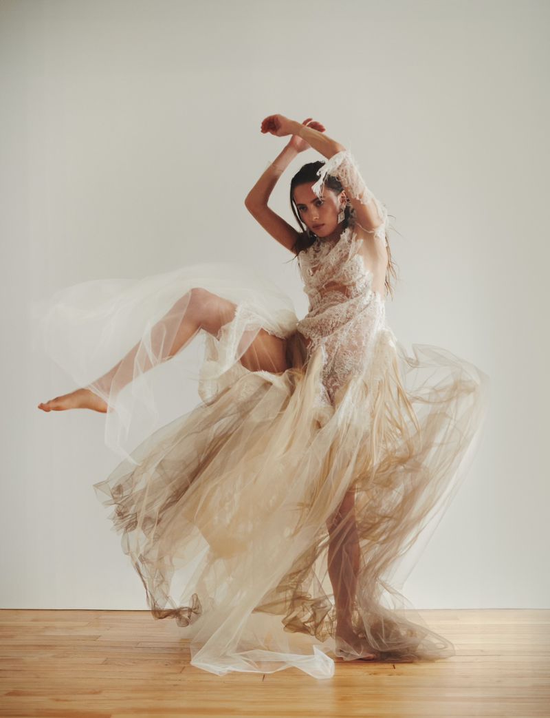 Woman in motion wearing lace dress with tulle overlay in photo studio setting with neutral background and light wood floor.