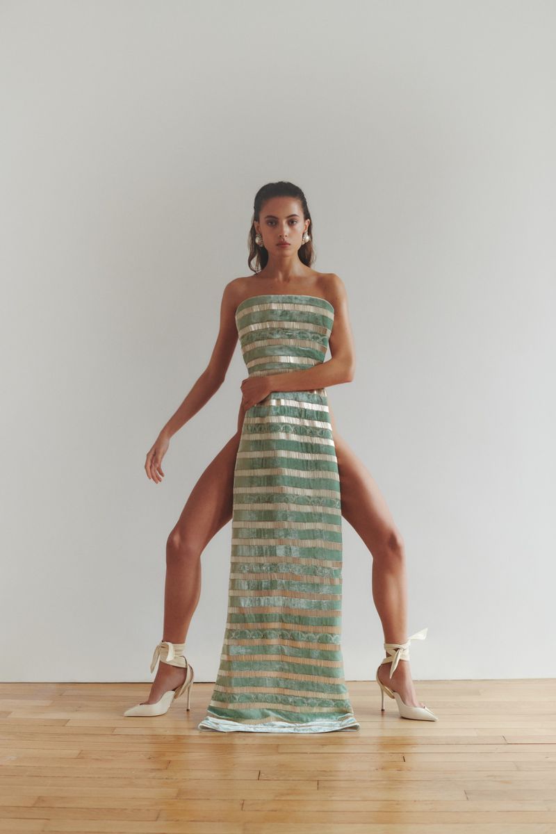 Fashion portrait of a woman posing in an indoor studio, wearing a strapless green and gold striped dress and white high-heeled shoes.