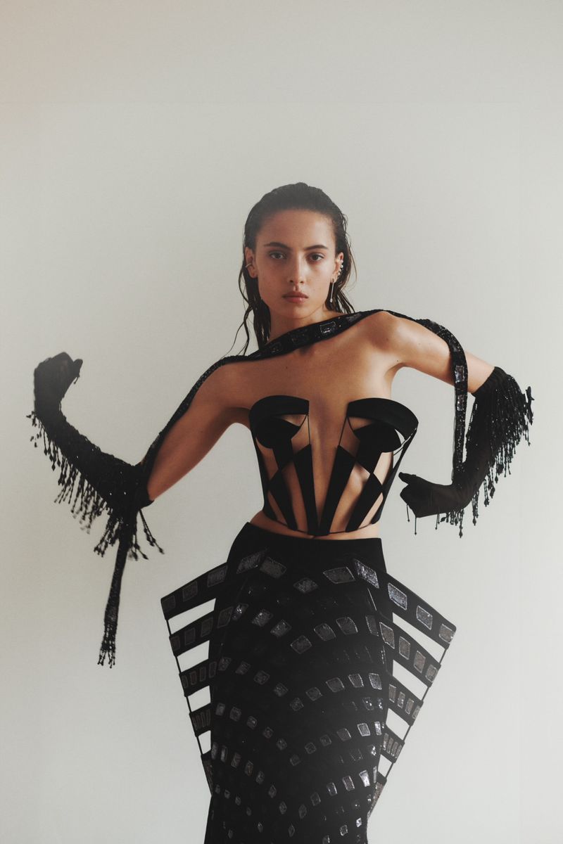 A woman poses in a black couture dress with beaded gloves against a white backdrop, presenting a high-fashion aesthetic and sleek styling.
