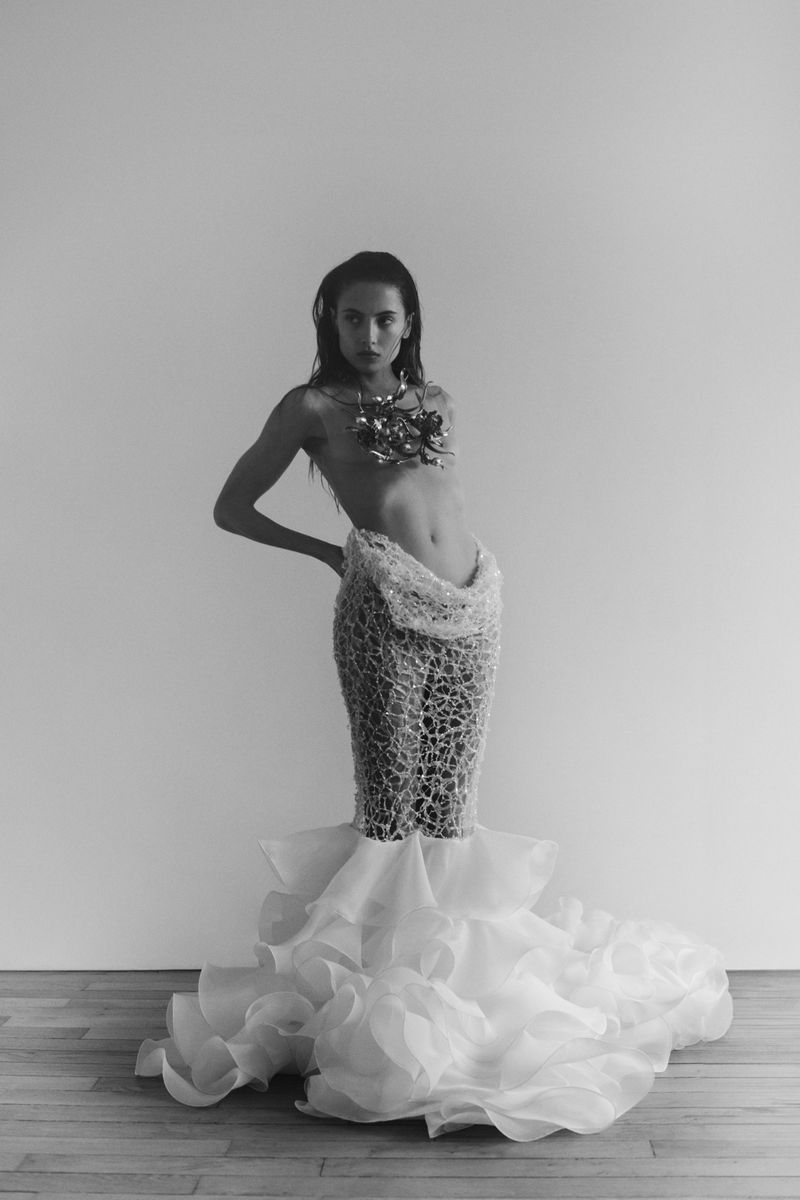 A woman models a couture gown with a structural bodice and cascading ruffled skirt in a black and white studio fashion photograph.