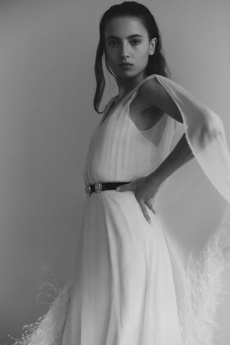 Woman poses in an elegant white dress with a dark belt and sheer shawl in a grayscale, indoor portrait with soft lighting.