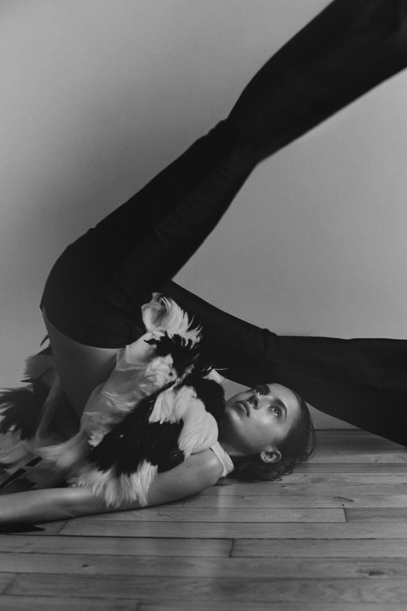 Monochrome fashion shot of a woman in black pants and a feathered top lying on a wood floor with her legs raised.