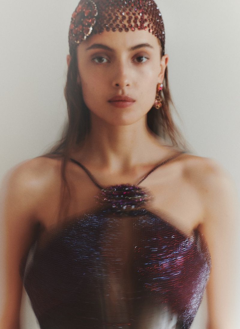 A woman with long dark hair poses in a shimmery burgundy halter top and jeweled cap against a neutral background in a beauty shot.