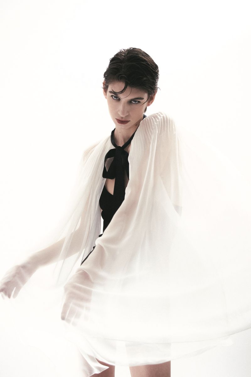 Fashion portrait of a female model with short hair, wearing a black top and a flowing sheer white cape against a bright white backdrop.