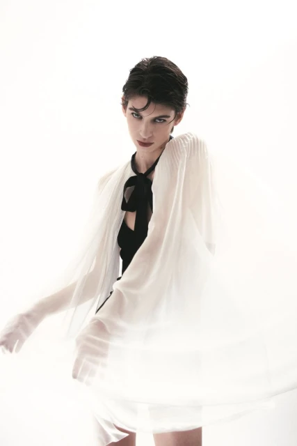 Fashion portrait of a female model with short hair, wearing a black top and a flowing sheer white cape against a bright white backdrop.