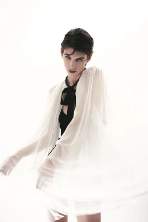 Fashion portrait of a female model with short hair, wearing a black top and a flowing sheer white cape against a bright white backdrop.