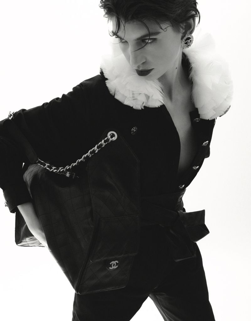 Black and white portrait of a young adult woman wearing a black jacket with white fur collar and carrying a black handbag with silver chain.
