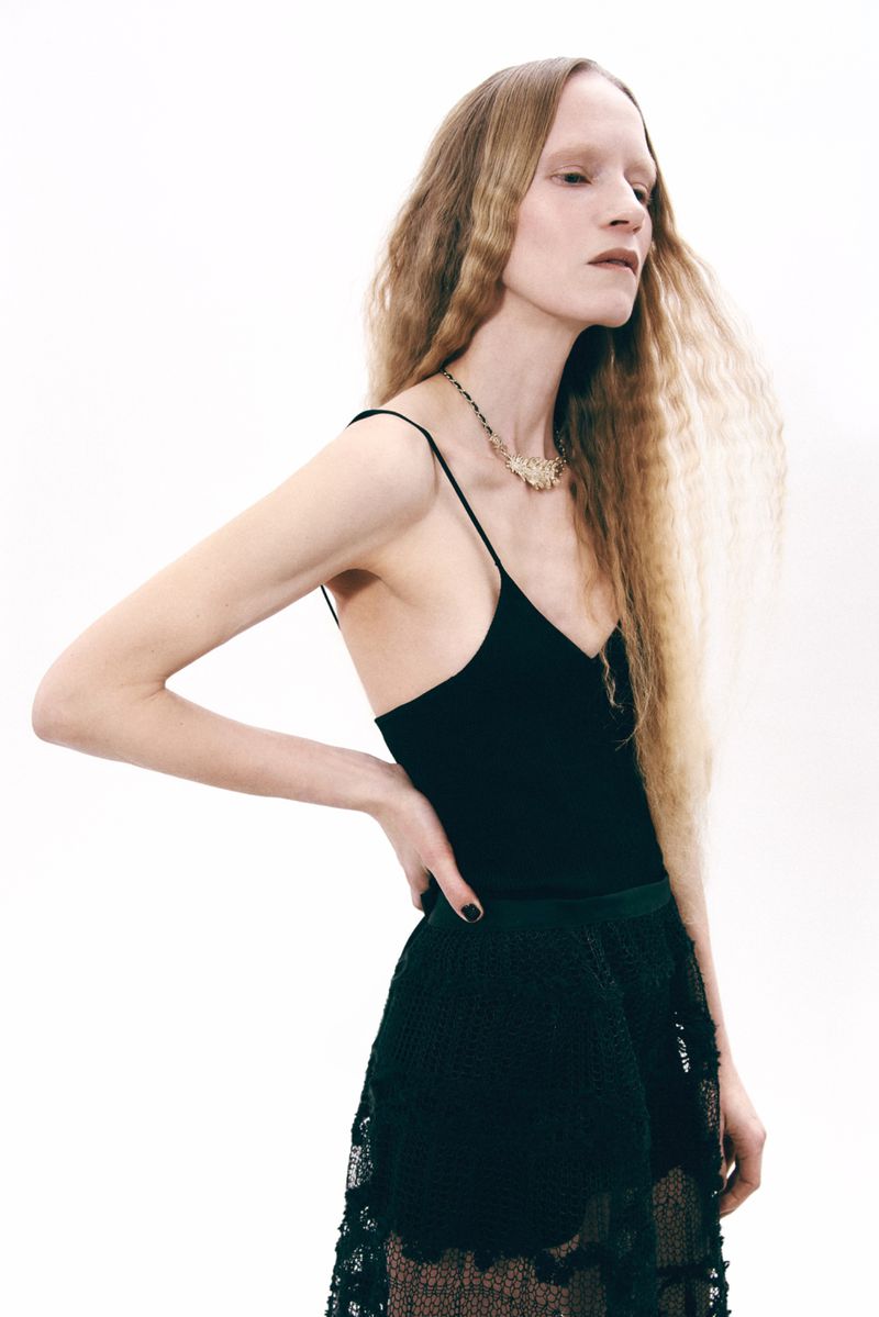 Elegant woman with long wavy hair poses in a black top and textured skirt against a stark white backdrop, fashion forward and chic.