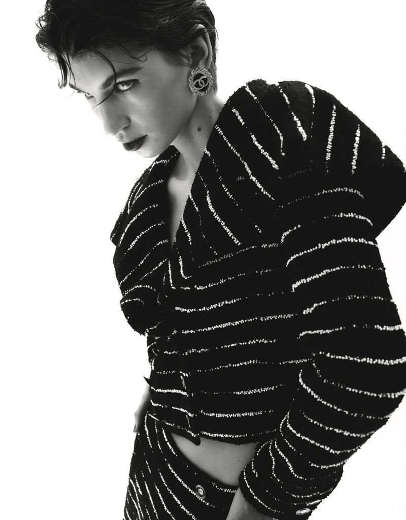 A fashionable adult woman in a patterned black and white suit poses in a studio setting, showcasing beauty makeup and stylish earrings.