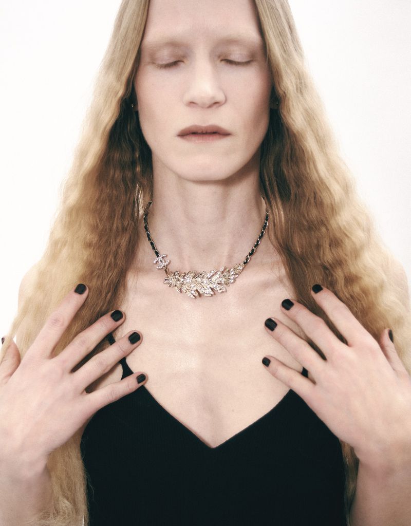 Adult female with eyes closed poses wearing a crystal necklace in front of a white background. Black manicure and black top.