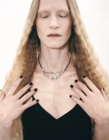 Adult female with eyes closed poses wearing a crystal necklace in front of a white background. Black manicure and black top.