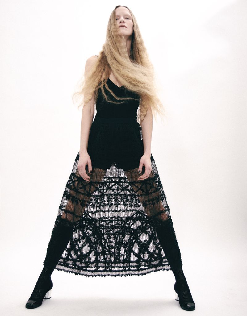 A striking fashion shot of a slender blonde woman in a black bodysuit and lace skirt, standing against a bright white backdrop with flowing hair.