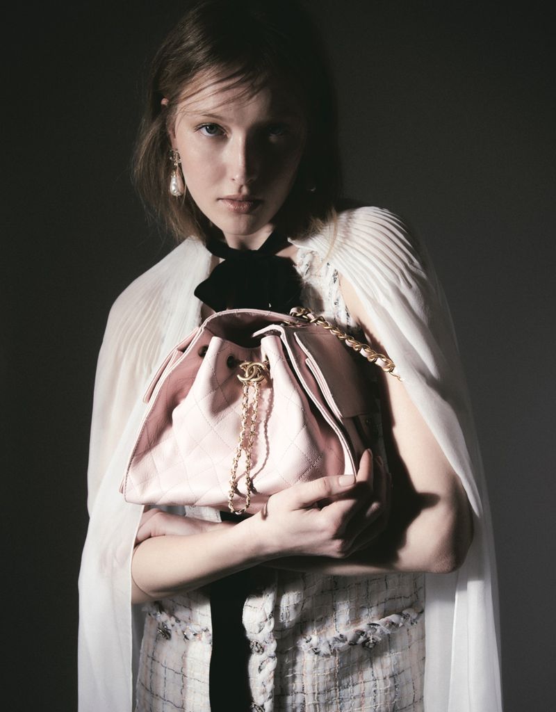 Woman poses with a pink Chanel handbag. She's wearing a white cape and dress against a dark gray background. Fashion editorial.