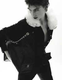 Model in black top with white fluffy collar holding a black quilted chain handbag against a white background