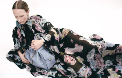 Model in a black patterned dress lying down and holding a light blue shoulder bag against a white background