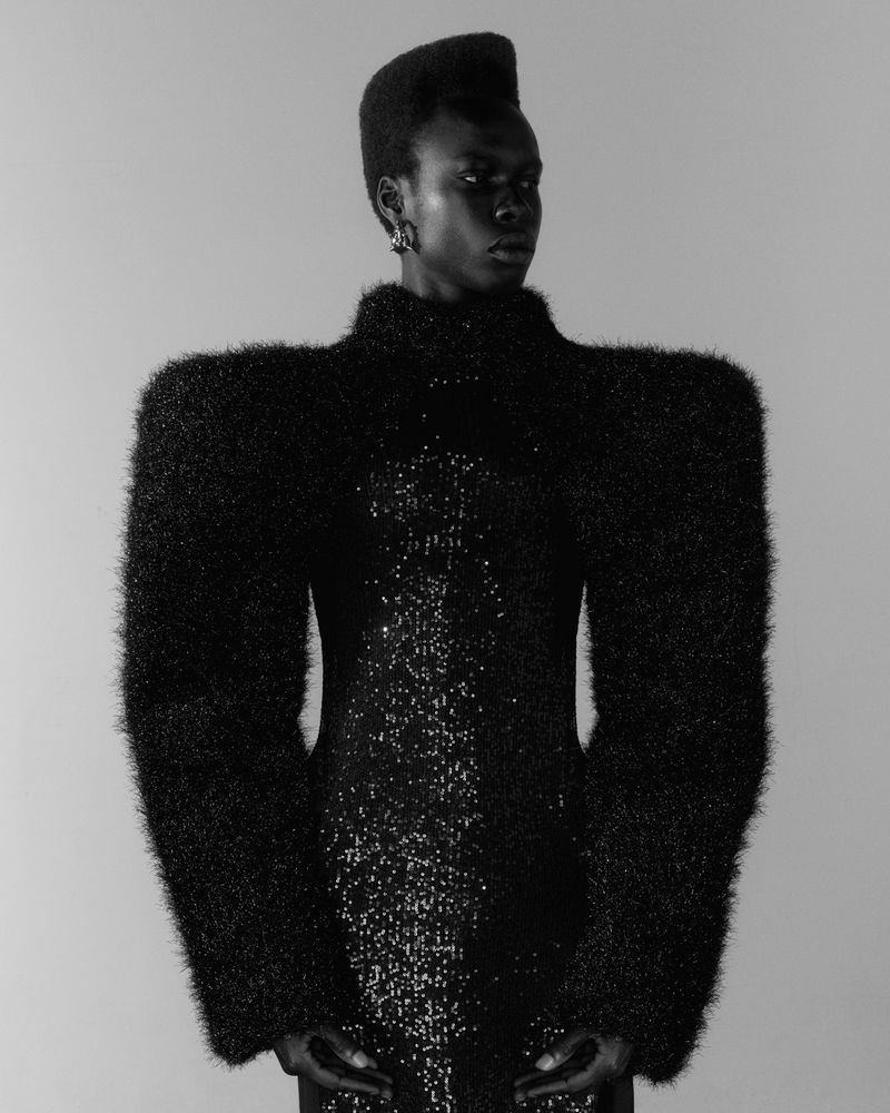 A striking portrait in black and white features a Black woman in a black dress with textured sleeves against a plain background.