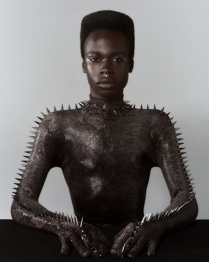 Fashion portrait of a Black adult female wearing a studded top sitting indoors with a light-grey background in a photo studio.