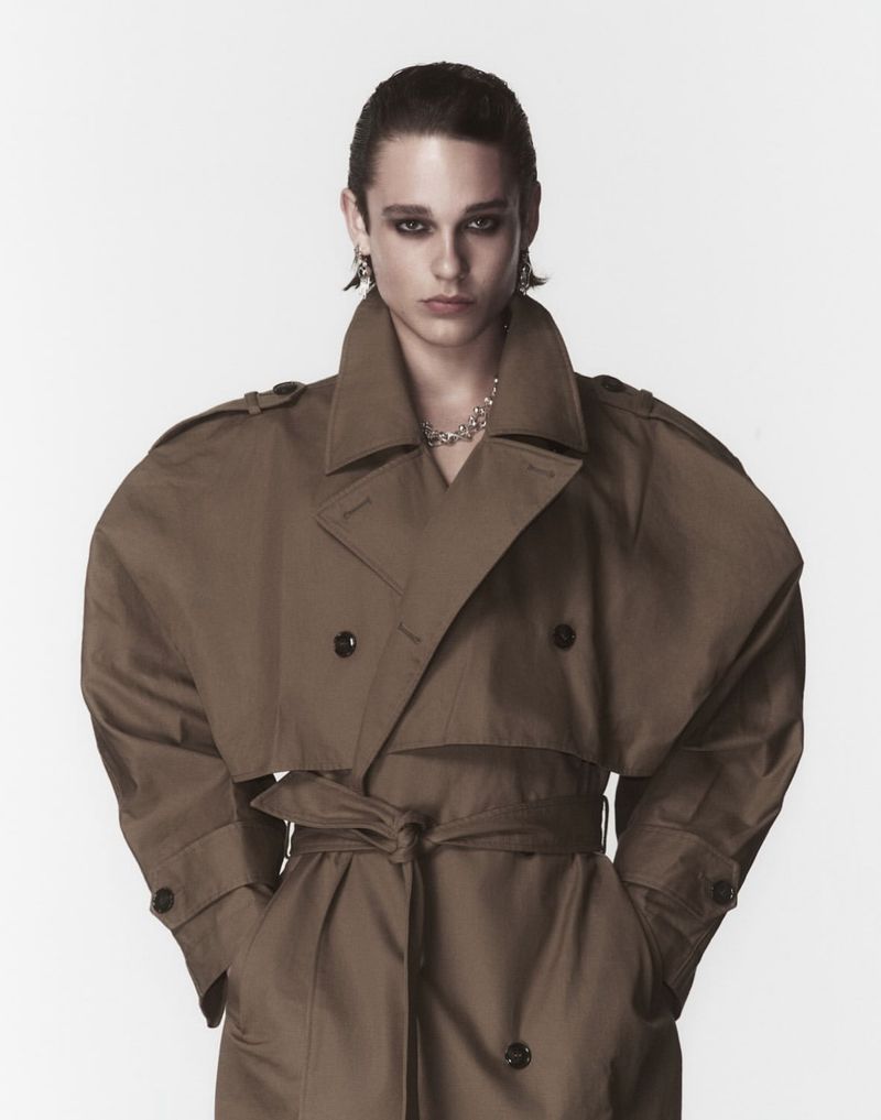 An androgynous model in a studio shot, wearing a trench coat, heavy eye makeup, jewelry, and posing against a light background.