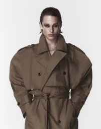Model wearing an oversized brown trench coat with dark eye makeup against a seamless white background.