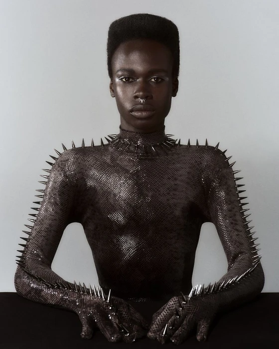 Portrait of a Black model in a dark, studded textured garment against a plain grey backdrop