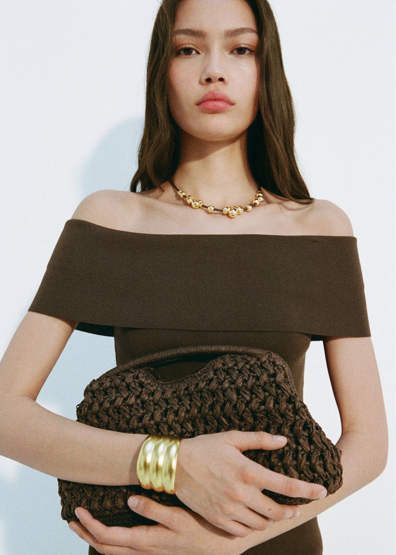 Portrait of a woman wearing a brown off-the-shoulder dress, gold necklace and bracelet, and holding a brown woven handbag.
