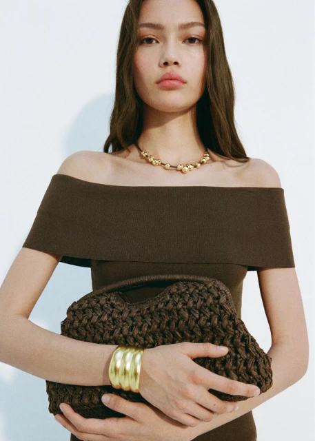Portrait of a woman wearing a brown off-the-shoulder dress, gold necklace and bracelet, and holding a brown woven handbag.