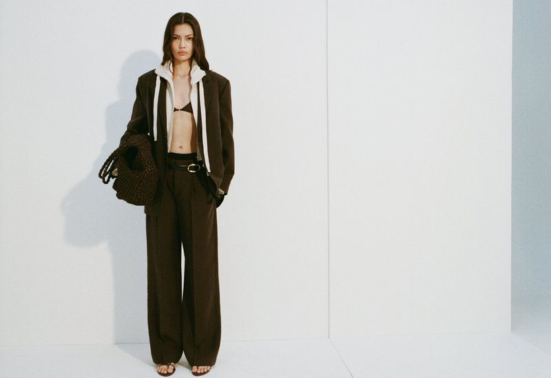 Full-length shot of a brunette woman in a brown blazer and trousers, accessorized with a knit bag, standing indoors against a white background.