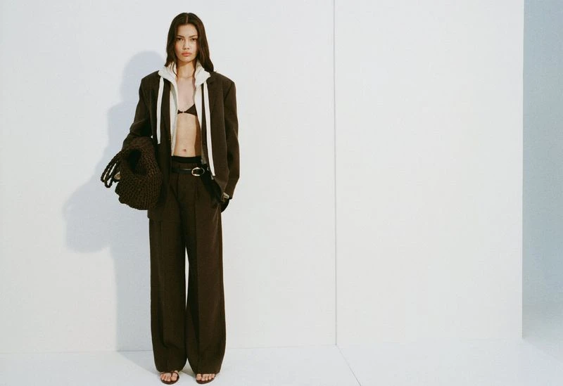 Full-length shot of a brunette woman in a brown blazer and trousers, accessorized with a knit bag, standing indoors against a white background.