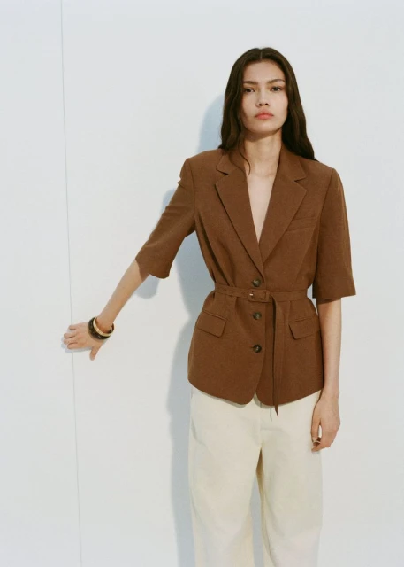 Brunette woman wearing a brown, short-sleeved, belted blazer and light pants posing in front of a bright, neutral background.
