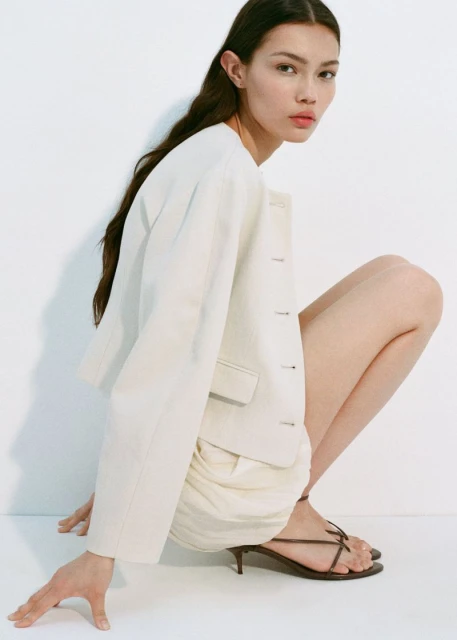 Woman in a crouched pose wearing a cream-colored blazer with matching shorts and heels against a white studio backdrop.