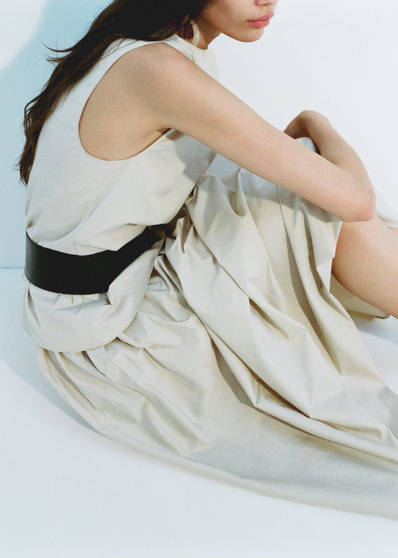 Woman in a light beige dress and wide black belt is sitting in a photo studio, posing. Part of her face and torso are visible.